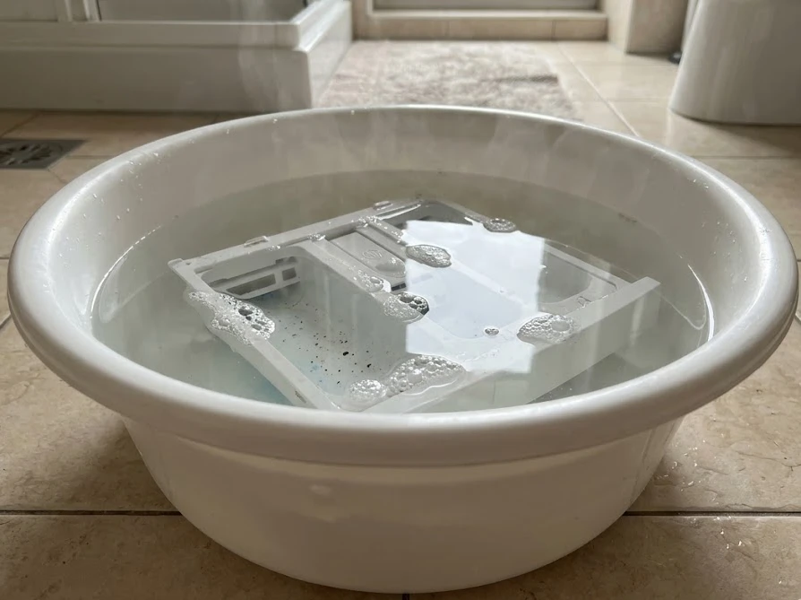 洗面器に溜めたぬるま湯に、取り外した洗剤投入口のパーツを浸して洗う様子